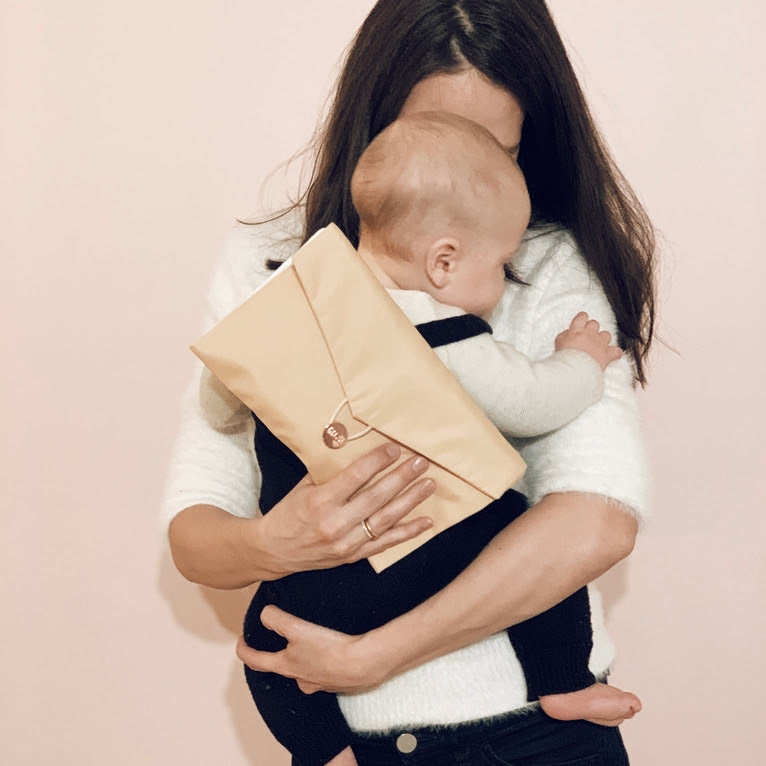 A woman cradles a baby against a light background, showcasing the elegant versatility of the Daily Daisy Swaddle Bag by Sonia's Vokalized Kreations, which doubles as a chic beige clutch resting on the baby's back.