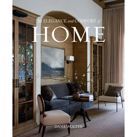 A cozy living room features Sonia's Vokalized Kreations' Boho Self-Care Home Gift Box, dark sofa, light armchair, glass cabinets, a large painting, and neutral decor. Overlaid: "The Elegance and Comfort of Home" by Dana Wolter.