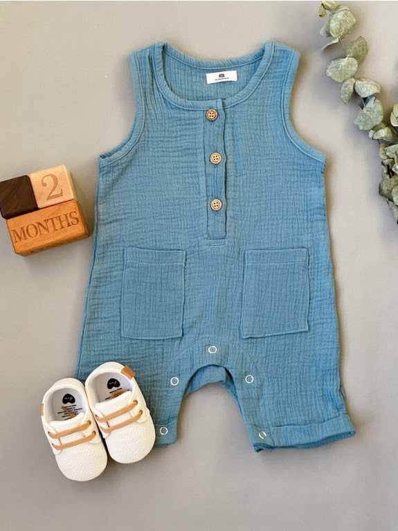 Sonia's Vokalized Kreations' New Baby Gift Box for Mom & Baby features a blue organic sleeveless jumpsuit with buttons and pockets, white baby shoes, "2 months" blocks, and green foliage on a gray background—an ideal gift.