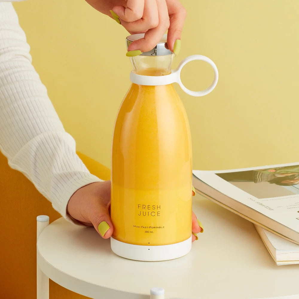 A hand holds a sonia's-vokalized-kreations Portable Blender Bottle, filled with orange liquid and labeled "Fresh Juice," resting on a small round table next to a book.