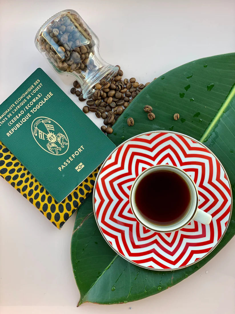An elegantly arranged display of a Togo passport, Itadi Coffee beans from family-owned farms, and an earthy-flavored coffee in a red and white patterned saucer rests on large green leaves, beautifully crafted by sonia's-vokalized-kreations.