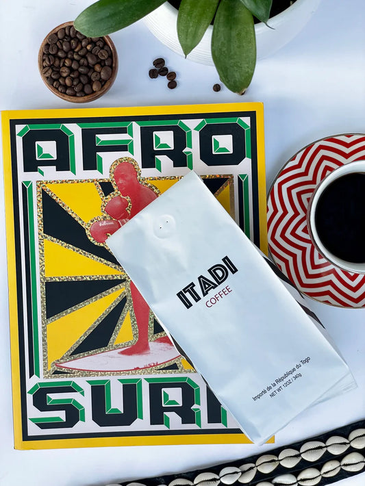 A bag of Itadi Coffee by sonia's-vokalized-kreations, celebrated for its earthy flavor, rests upon an Afro Surrealism book alongside a filled cup on a red-patterned saucer. Encircled by beans from family-owned farms and a lush plant, it epitomizes indulgence and culture.