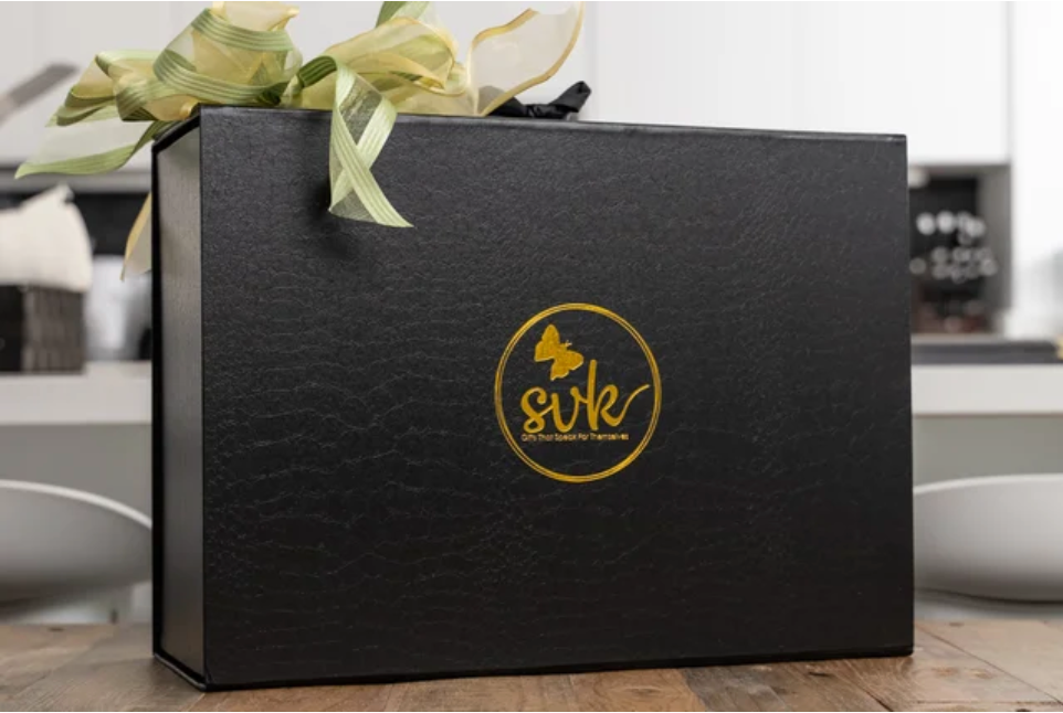 A black gift box with a gold sonia's-vokalized-kreations logo and ribbon rests elegantly on wood, capturing the essence of the trending Surprise Me Kreation.