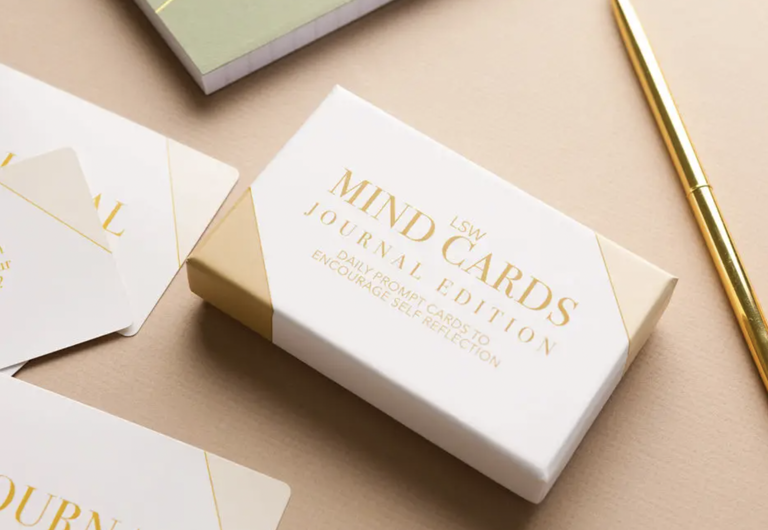 A box of "Mind Cards: Journal Edition" by sonia's-vokalized-kreations sits on a beige surface, accompanied by a gold pen and scattered cards filled with insightful journal prompts for your self-discovery journey.