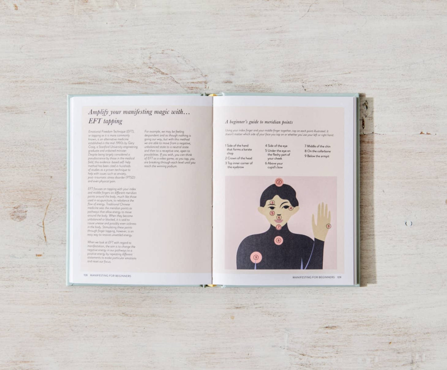 On a light surface, an open "Manifesting For Beginners" by Victoria Jackson (sonia's-vokalized-kreations) reveals two pages: the left filled with insights on EFT Tapping for mindset shifts, and the right displaying an illustration of a person with labeled positive-energy points.