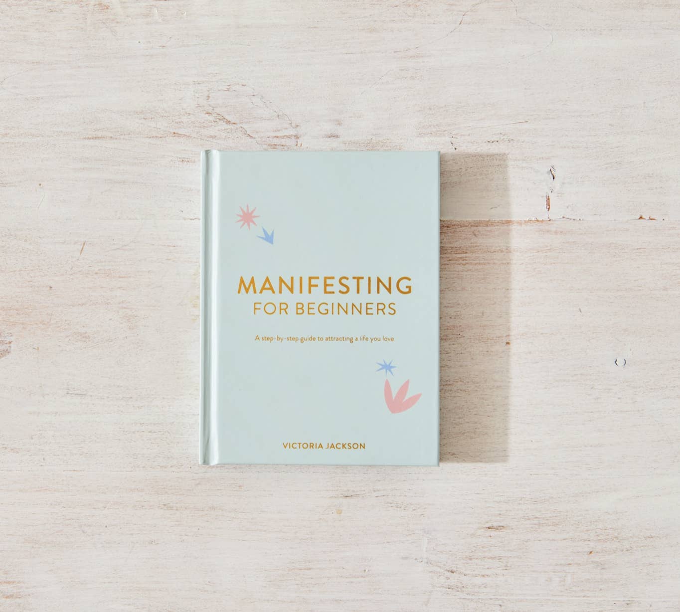 The book "Manifesting For Beginners" by Victoria Jackson from sonia's-vokalized-kreations, offering insights on positive energy and mindset shifts, rests beautifully on a light wooden surface.