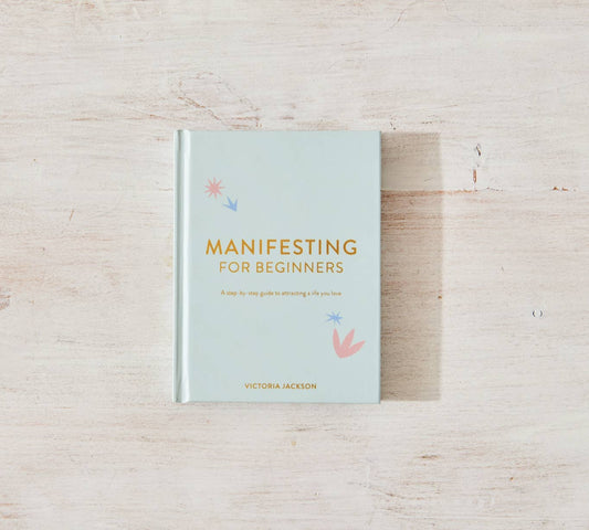 The book "Manifesting For Beginners" by Victoria Jackson from sonia's-vokalized-kreations, offering insights on positive energy and mindset shifts, rests beautifully on a light wooden surface.