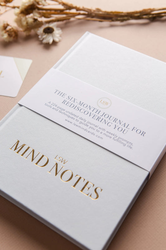 A white "Mind Notes Journal" by sonia's-vokalized-kreations, adorned with gold lettering, rests on a surface. A band highlights six months of life fulfillment guidance. Dried flowers in the background enhance its charm, inviting a gratitude practice journey.