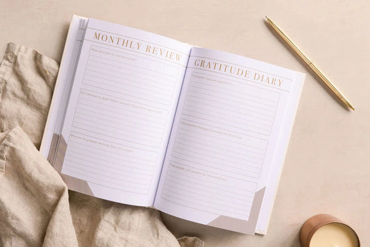 The Morning Notes Journal by sonia's-vokalized-kreations rests on a beige cloth, featuring a monthly review and prompts to encourage positivity. A gold pen lies on the right, ready to record your daily well-being journey.