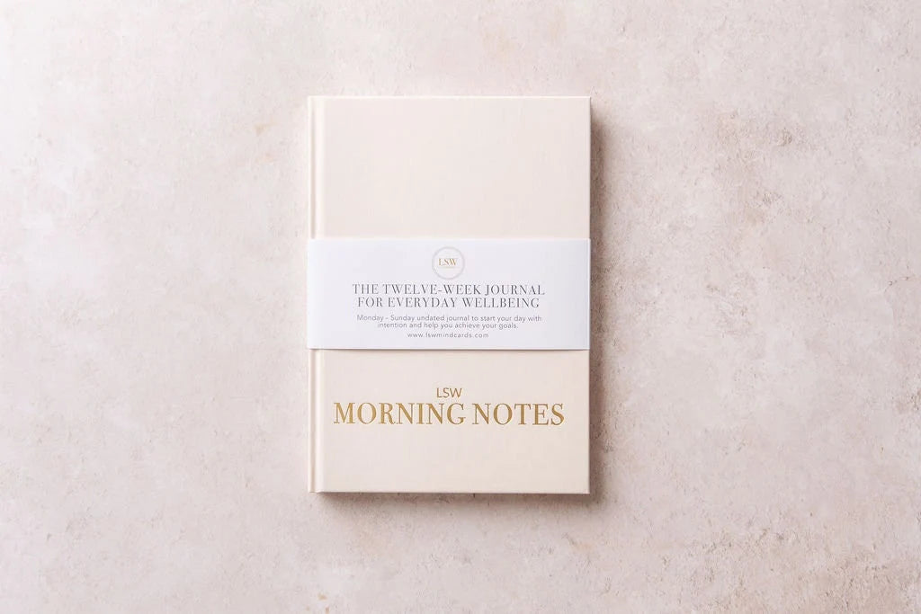 A beige hardback "Morning Notes Journal" from sonia's-vokalized-kreations rests on a textured surface. Wrapped in a paper band, it reads: "The Twelve-Week Daily Well-being Journal for Everyday Wellbeing," inspiring healthy habits and positivity.