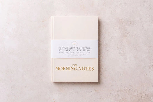 A beige hardback "Morning Notes Journal" from sonia's-vokalized-kreations rests on a textured surface. Wrapped in a paper band, it reads: "The Twelve-Week Daily Well-being Journal for Everyday Wellbeing," inspiring healthy habits and positivity.