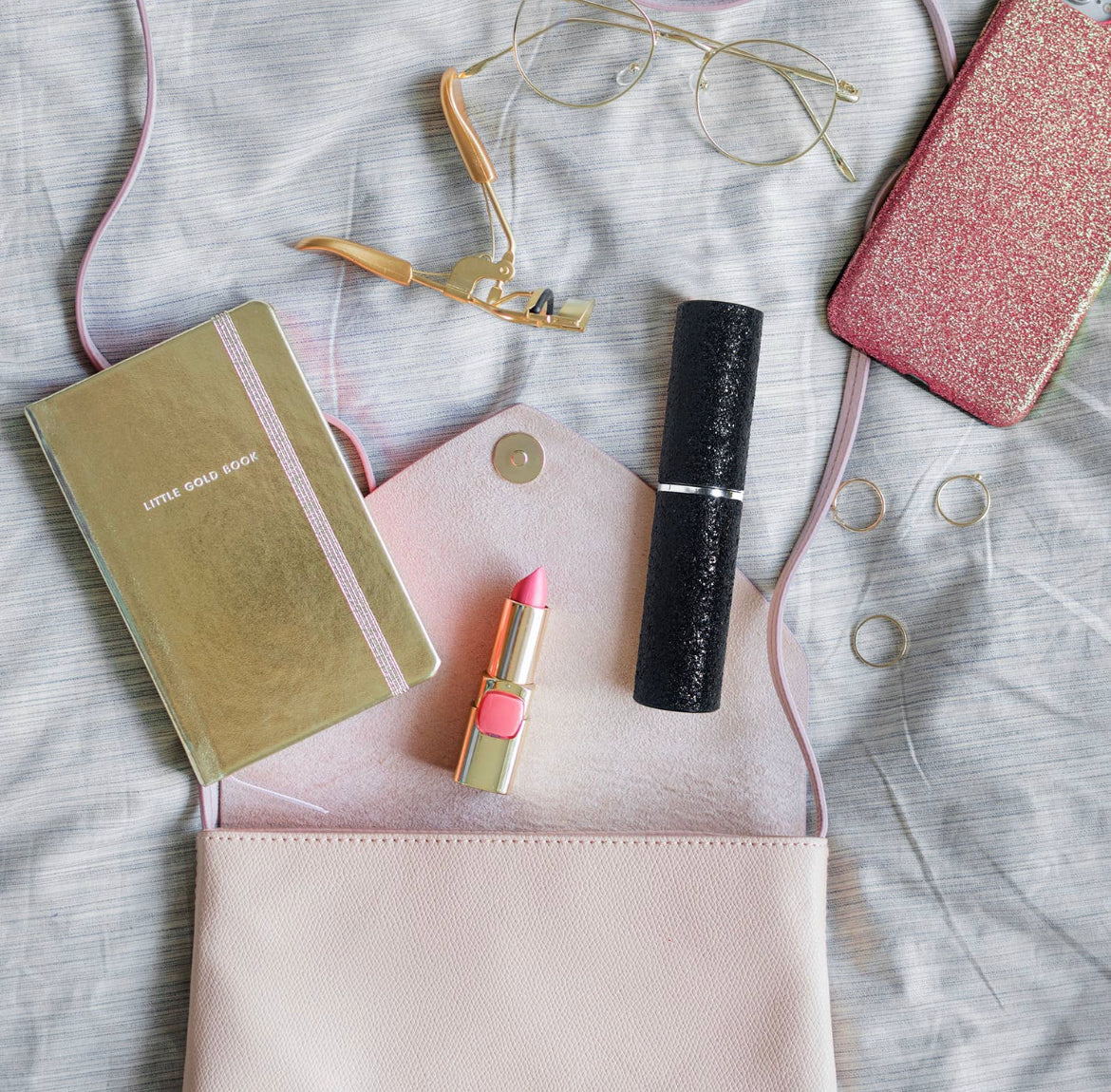 An open pink purse holds a notebook, glasses, lipstick, mascara, and gold rings. Beside it on a gray fabric background are a sparkly pink phone case and rechargeable pen, perfectly complementing sonia's-vokalized-kreations' Mini Stun Gun for the modern women's safety brand.