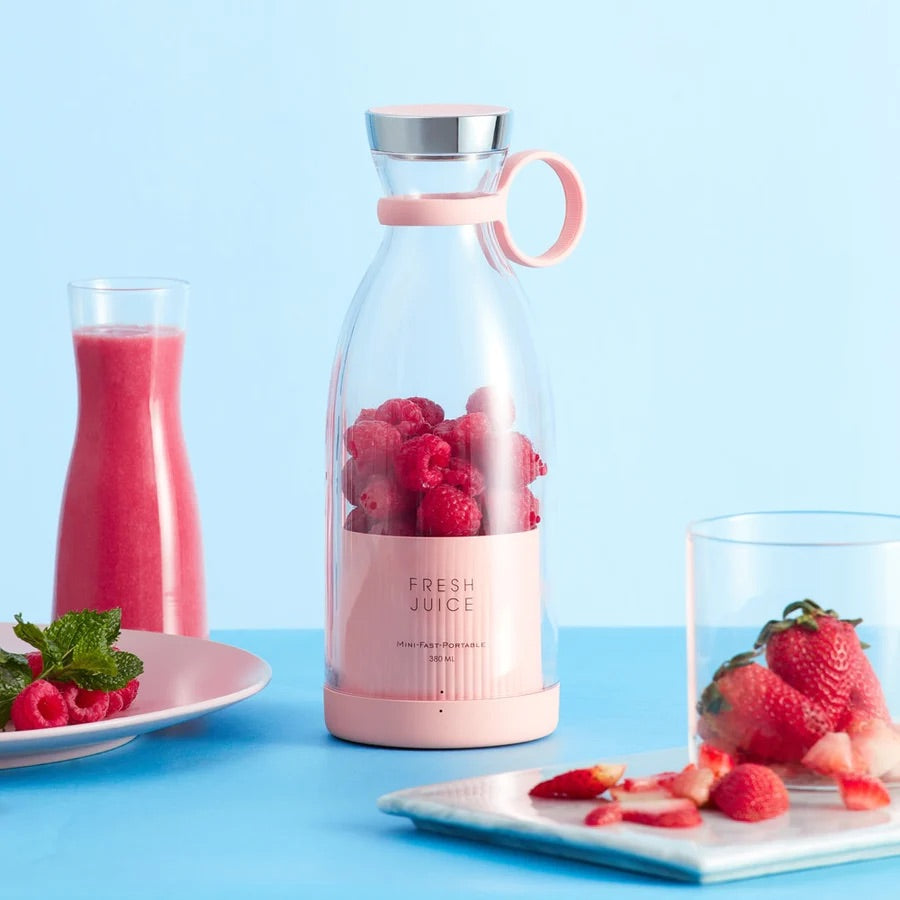 Sonia's Vokalized Kreations' BPA-free Portable Blender Bottle in pink, ideal for on-the-go smoothies with wireless charging, is filled with raspberries. Nearby, a glass of vibrant pink smoothie and a plate of strawberries elegantly sit against a blue background.