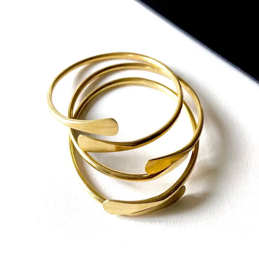 Three slender brass coil bangles by Sonia's Vokalized Kreations, with overlapping curved ends, are stacked on a white surface.