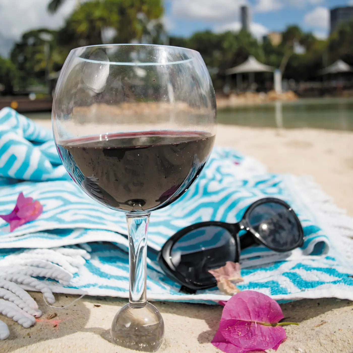 The Sonia's Vokalized Kreations Floating Wine Glass for Beach, Pool & Outdoors sits on a beach towel with sunglasses and pink flower petals on sand, near water with trees and buildings in the background.