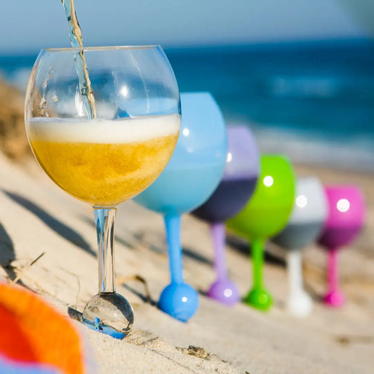 A drink is poured into a Sonia's Vokalized Kreations Floating Wine Glass for Beach, Pool & Outdoors (Reusable Acrylic) with a round base on sand, beside four colorful Beach Glasses, ocean in the background.
