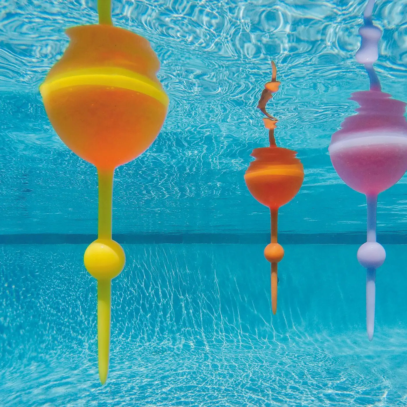 Three colorful pool toys float beneath the water’s surface next to a Sonia's Vokalized Kreations Floating Wine Glass for Beach, Pool & Outdoors – Reusable Acrylic in a bright blue swimming pool.