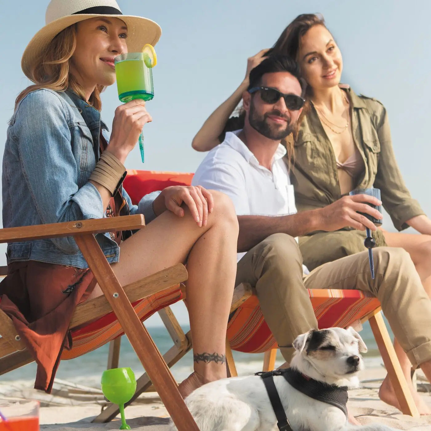 Three people relax on beach chairs, sipping drinks from Sonia's Vokalized Kreations Floating Wine Glasses. A small white dog with a harness lies on the sand nearby as they enjoy a sunny day by the water.