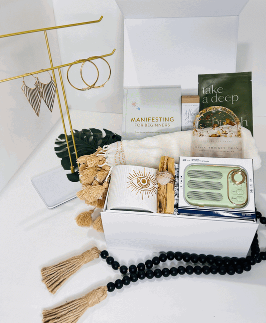 The "Manifestation Woman!" gift box by Sonia's Vokalized Kreations features a tasseled blanket, ceramic mug, green radio, tea packets, "Manifesting for Beginners" book, gold jewelry stand with earrings, black beaded tassel garland, and aloe vera oils.
