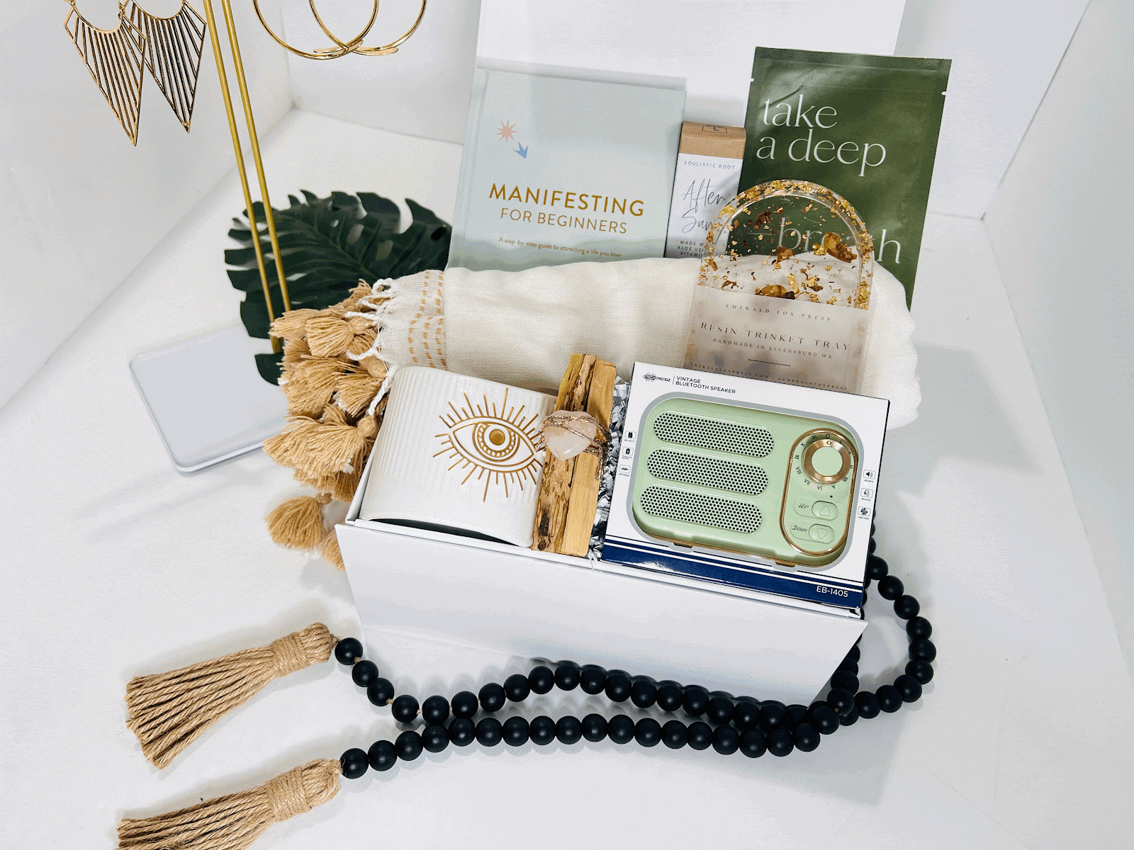 A white gift box from Sonia's Vokalized Kreations features the book "Manifestation Woman!", a green speaker, an eye design mug, journal, beads, towel, relaxing essential oils, and a sage stick.