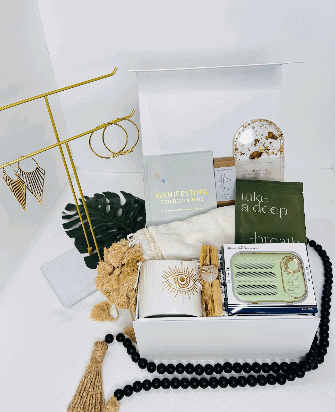 The Manifestation Woman! gift box by Sonia's Vokalized Kreations includes a book, mug, incense holder, tea bag, towel, beaded necklace, earrings on a stand, sunburn relief essentials, and other wellness items arranged neatly.