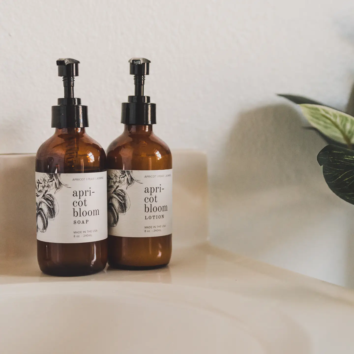 Two amber pump bottles from Sonia's Vokalized Kreations’ Boho Self-Care Home Gift Box sit on a bathroom sink beside a green plant, setting a tranquil, spa-like mood.