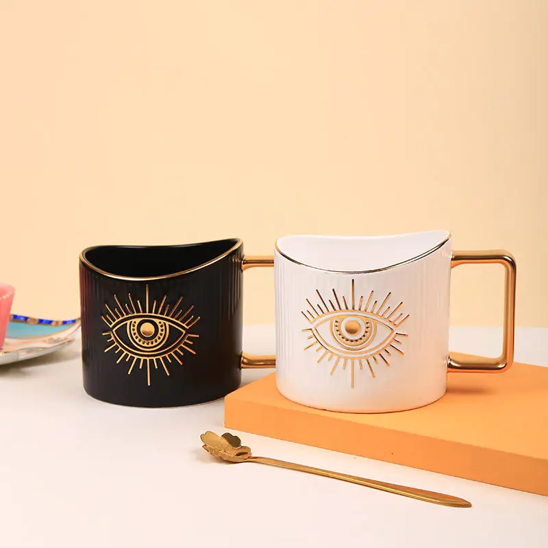 Two Manifestation Woman! ceramic mugs by Sonia's Vokalized Kreations—one black, one white, both with gold eye designs and handles—rest beside an orange book and gold spoon, perfect for sipping tea after applying sunburn relief.