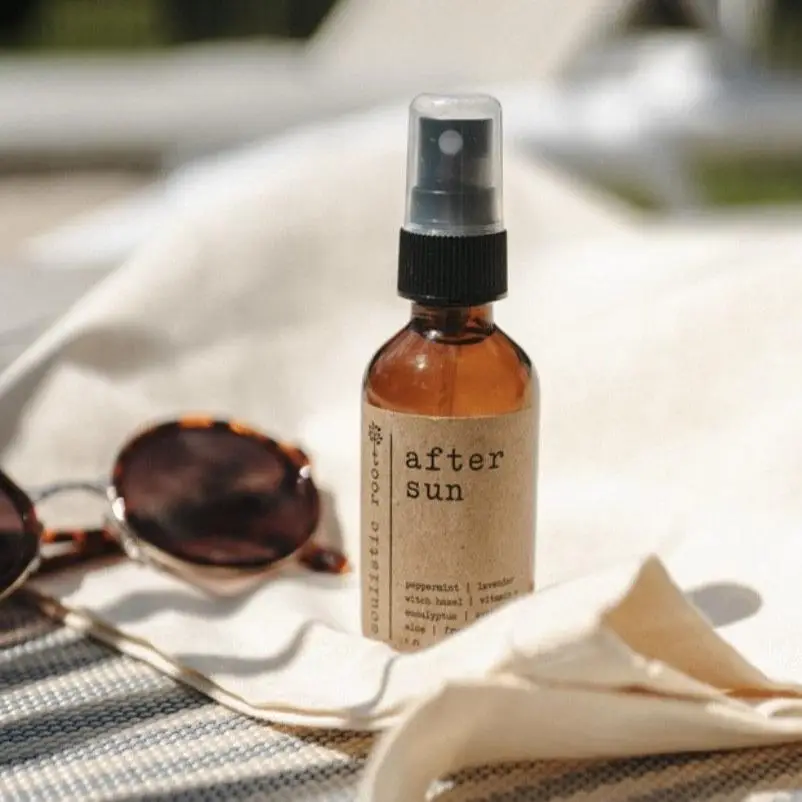A brown glass spray bottle labeled "Manifestation Woman!" by Sonia's Vokalized Kreations rests on a beige cloth beside tortoiseshell sunglasses, offering gentle sunburn relief with soothing essential oils.