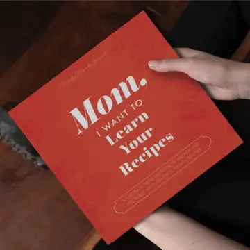 Someone holds the red "Mom, I Want To Learn Your Recipes" book by Sonia's Vokalized Kreations on their lap, ready to preserve treasured family recipes for future generations.