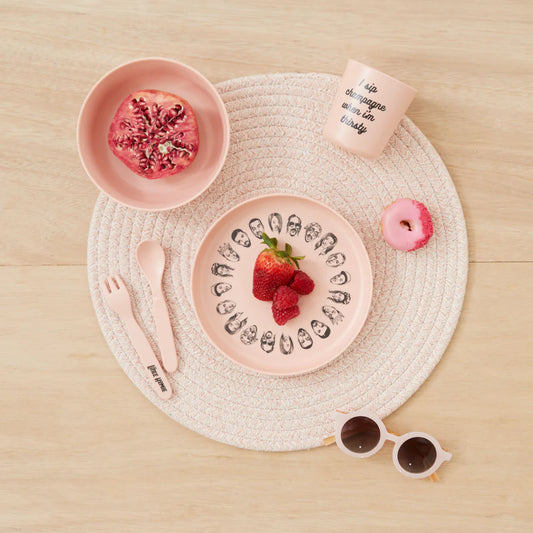 Sonia's Vokalized Kreations presents the Baby Bamboo Dinnerware Set: a pink, eco-friendly dishware arrangement featuring bowls with pomegranate halves and plates of raspberries. The set includes a text-printed cup, a small pink donut, wooden utensils, and sunglasses stylishly resting on a placemat.